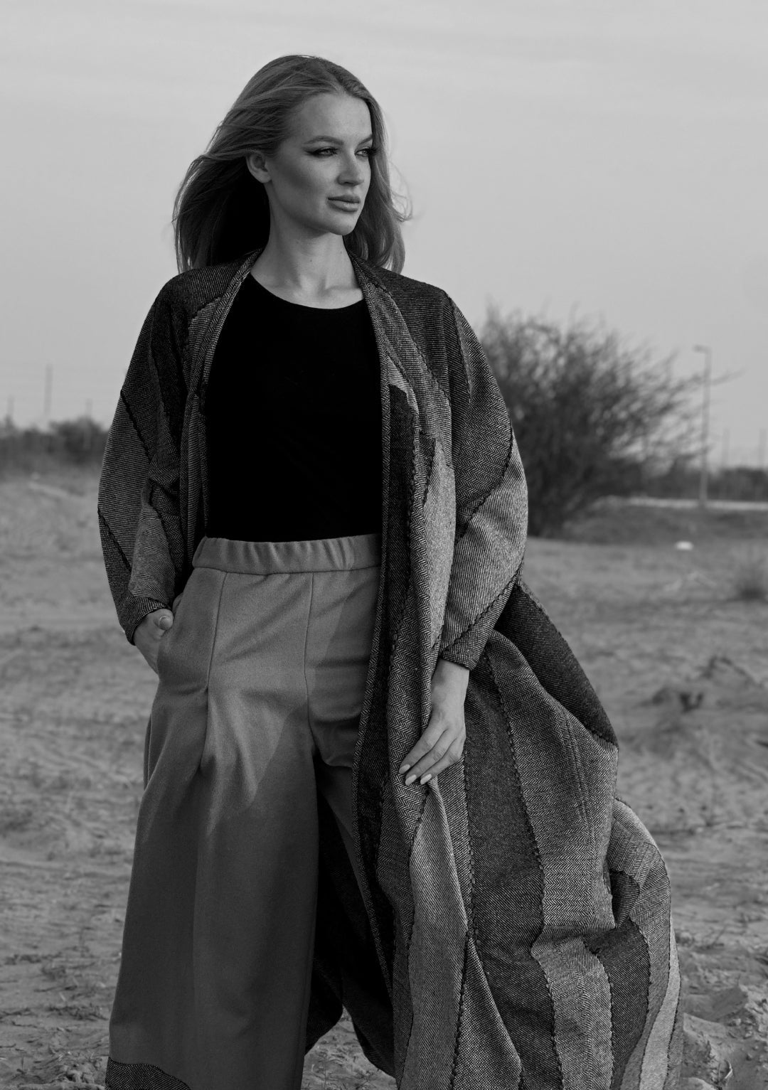Woman standing outdoors in a field wearing a winter woolen abayas, black top, and light pants contrast with an abaya.